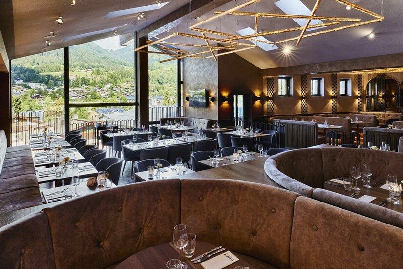 Moderner Restaurant-Innenraum mit gepolsterten Sitzbänken, Tischen und großer Fensterfront mit Bergblick.