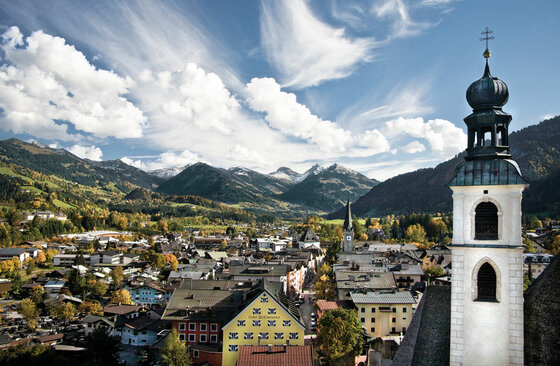 Blick über die Dächer von Kitzbühel mit Kirchturm im Vordergrund und Bergen im Hintergrund.