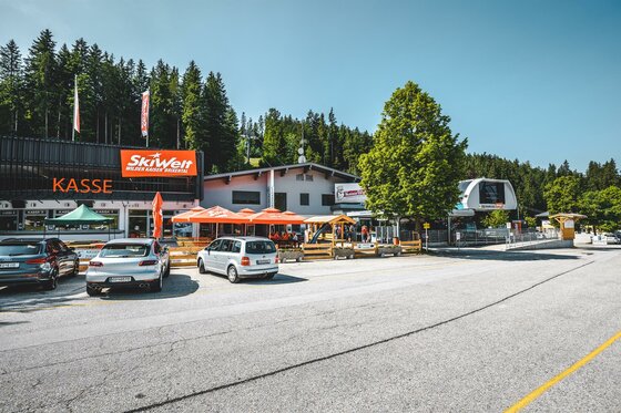 Talstation Bergbahn Scheffau 2 Eine belebte Bergstation mit einem Kassenbereich und zahlreichen Fahrzeugen. Die Umgebung ist grün und von Bäumen umgeben, ideal für Outdoor-Aktivitäten.