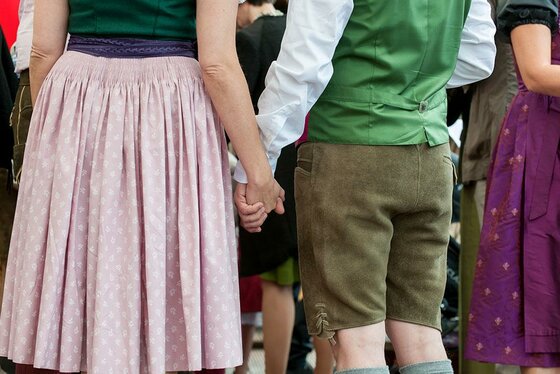 Sommer Ein Paar hält Händchen. Die Person links trägt einen Rock, während die Person rechts eine traditionelle Tracht mit kurzen Hosen trägt.