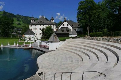 Kaps Ein malerisches Schloss mit einem grünen Umfeld und blauen Himmel. Im Vordergrund gibt es eine elegante Treppe, die zu einem Teich führt.