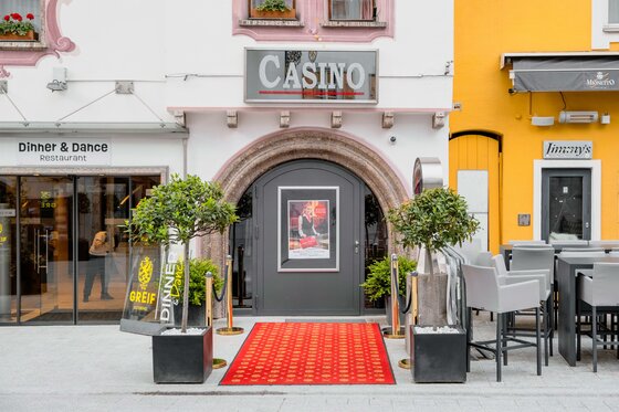 Casino2 Ein eleganter Casinoeingang mit einem roten Teppich und Pflanzen beidseitig. Daneben befindet sich ein Restaurant mit einem modernen Ambiente.