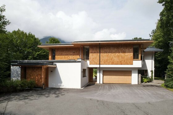 Ein modernes Haus mit Holzverkleidung und Garagentor in einer grünen Umgebung. Der Himmel ist teilweise bewölkt, und die Landschaft ist ruhig.