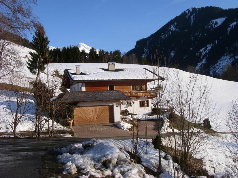 Alpines Wohnhaus mit Holzfassade und schneebedecktem Dach vor bewaldeten Bergen.
