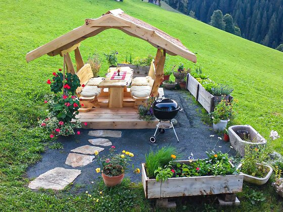 Überdachter Rastplatz am Bauernhof Überdachte Sitzgruppe aus hellem Holz mit Kugelgrill und Blumenkästen auf einer grünen Wiese.
