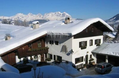 Landhaus Eva im Winter Schneebedecktes Landhaus mit dunkler Holzfassade vor einem Gebirgspanorama unter blauem Himmel.