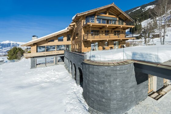 KitzChalet Jochberg im Winter Mehrstöckiges Holzchalet mit Steinfassade in einer verschneiten Berglandschaft unter blauem Himmel.