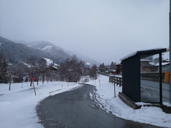 Bushaltestelle am Auweg Verschneite Straße mit Bushaltestelle und bewaldeten Bergen im Hintergrund.