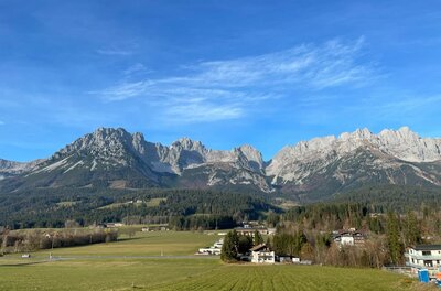 Panorama Wilder Kaiser Blick auf das Gebirgsmassiv des Wilden Kaisers über grüne Wiesen und vereinzelte Häuser unter blauem Himmel.