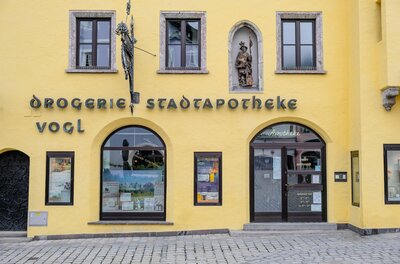 Stadtapotheke Vogl Gelbe Fassade der Stadtapotheke Vogl mit Rundbogenfenstern, einer Heiligenfigur und historischer Beschriftung.