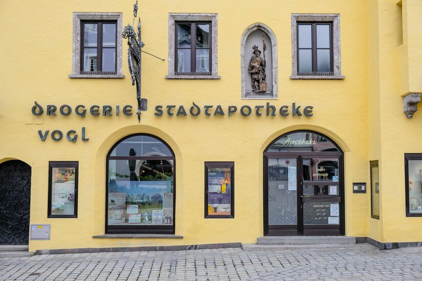 Gelbe Fassade der Stadtapotheke Vogl mit Rundbogenfenstern, einer Heiligenfigur und historischer Beschriftung.