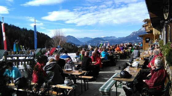 Eine belebte Terrasse mit vielen Gästen, die die Sonne genießen. Im Hintergrund sind Berge und ein klarer Himmel zu sehen.
