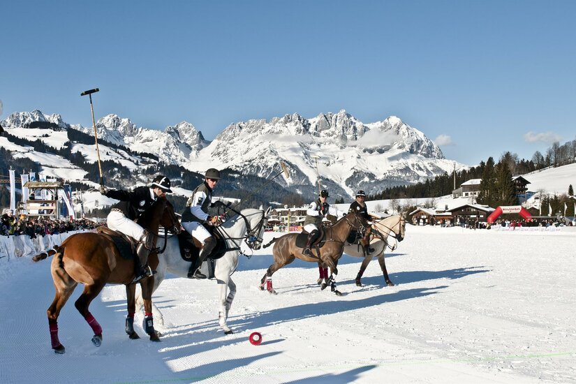 Vier Polospieler auf Pferden während eines Spiels auf einer Schneefläche vor einem verschneiten Bergmassiv.