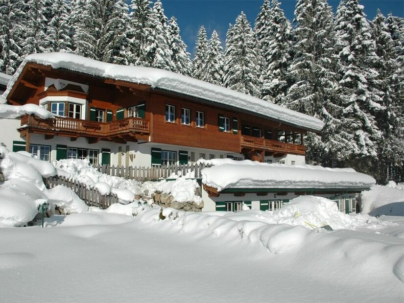 Verschneites Holzhaus mit Balkon vor einem Nadelwald im Winter.