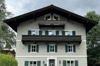 Vorderansicht der Villa Anna mit grauer Fassade, grünen Fensterläden und Holzbalkonen unter bewölktem Himmel.