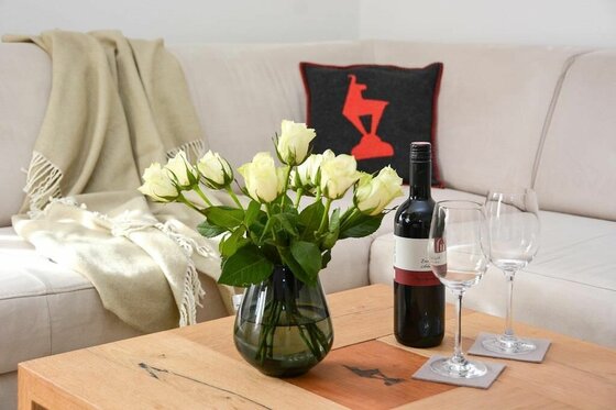 Weiße Rosen in einer Glasvase neben einer Weinflasche und zwei Gläsern auf einem Holztisch vor einem Sofa.