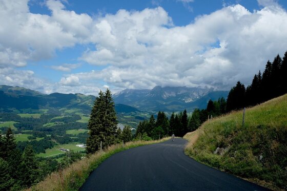 Asphaltierte Bergstraße am Kitzbüheler Horn mit Blick auf bewaldete Hänge und Berge unter bewölktem Himmel.