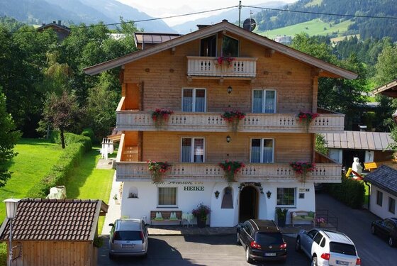 Mehrstöckiges Holzhaus mit Balkonen, Blumenschmuck und Parkplatz davor in alpiner Landschaft.