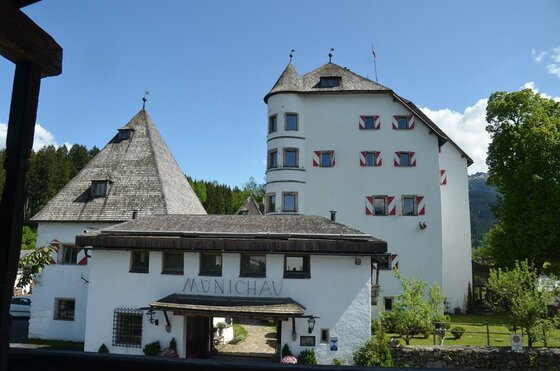 Hotel Schloss Münichau Weißes Schlossgebäude mit Rundturm, Holzschindeldächern und rot-weiß gestreiften Fensterläden unter blauem Himmel.