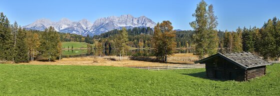 Blick über eine grüne Wiese und einen See auf das Gebirgsmassiv des Wilden Kaisers unter blauem Himmel.