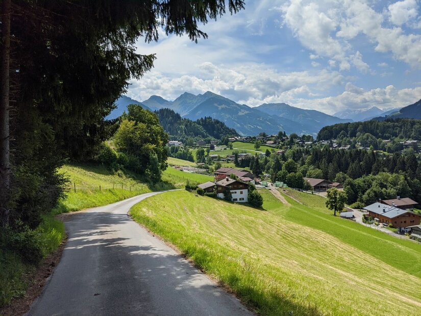 Asphaltierter Weg führt an grünen Wiesen und Häusern vorbei auf ein Bergpanorama zu.