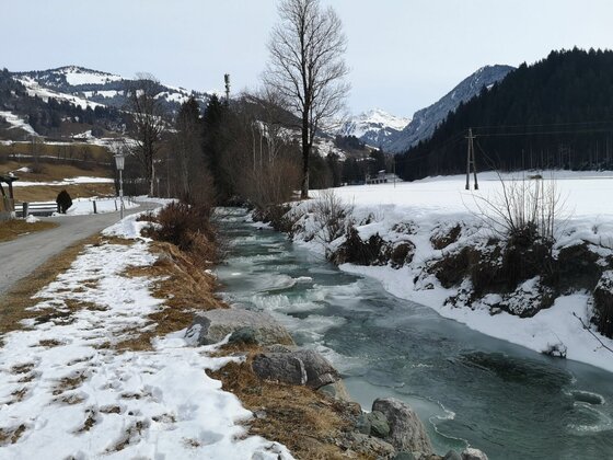 Ein teilweise zugefrorener Bachlauf in einer verschneiten Gebirgslandschaft mit angrenzendem Wanderweg.