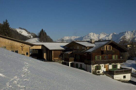 Alpiner Ferienhof aus Holz und Stein in einer verschneiten Berglandschaft unter blauem Himmel.