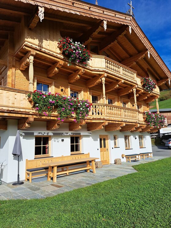 Ein traditionelles Holzhaus mit blühenden Blumenkästen. Es steht in einer grünen Umgebung und hat eine einladende Terrasse.