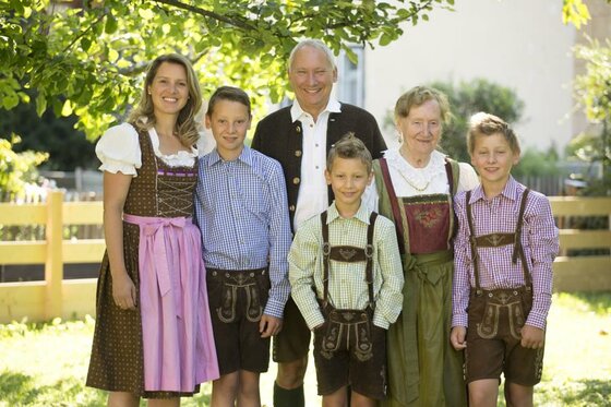 Sechs Personen in traditioneller Tracht stehen lächelnd in einem Garten vor einem Holzzaun.