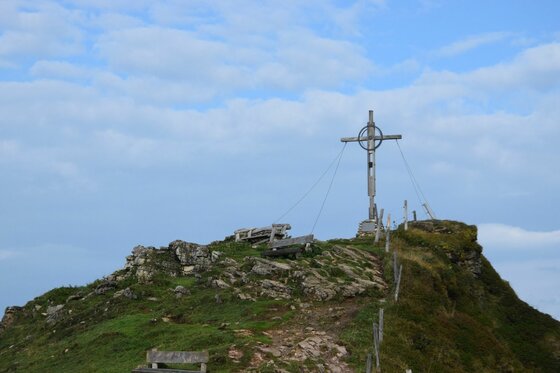 Ein hölzernes Gipfelkreuz auf einem felsigen, grasbewachsenen Berggipfel unter leicht bewölktem Himmel.