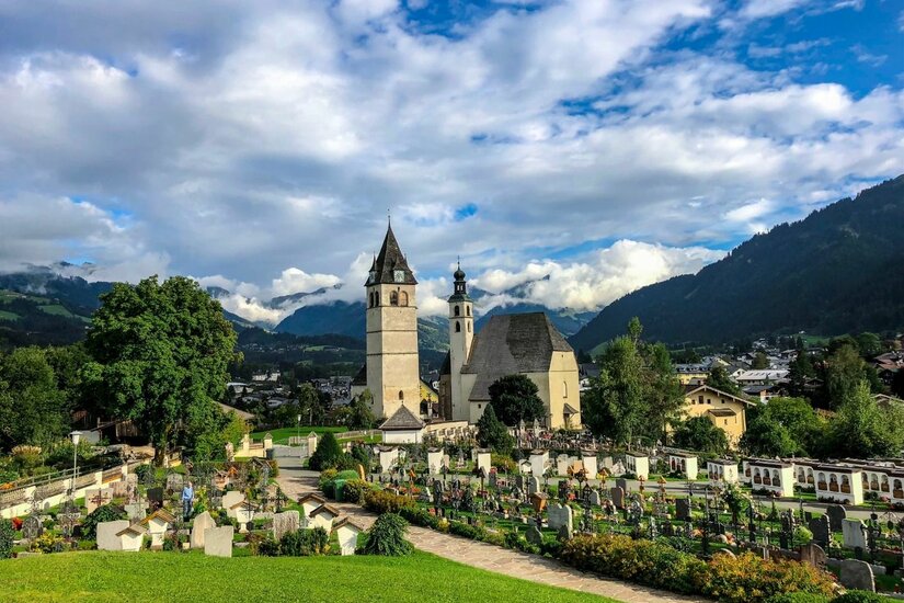 Blick auf eine Kirche mit zwei Türmen und einen angrenzenden Friedhof vor einer Bergkulisse.