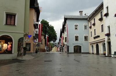 Gepflasterte Fußgängerzone in Kitzbühel mit bunten Häuserfassaden und rot-weißen Fahnen unter bewölktem Himmel.