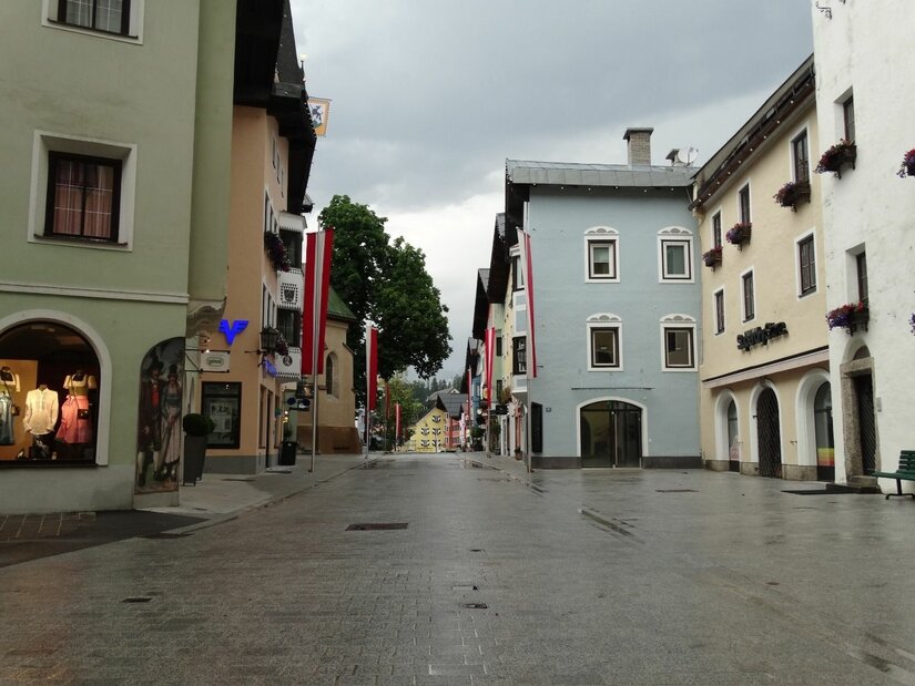 Gepflasterte Fußgängerzone in Kitzbühel mit bunten Häuserfassaden und rot-weißen Fahnen unter bewölktem Himmel.