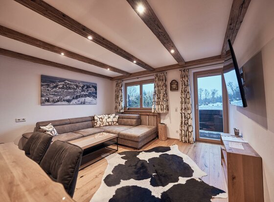 Wohnzimmer Ferienwohnung Hahnenkammblick Modernes Wohnzimmer mit Ecksofa, Holzbalkendecke, Kuhfellteppich und Bergbild an der Wand.