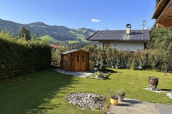 Gartenanlage Tannenheim Garten mit Rasenfläche, Holzschuppen und Wohnhaus vor bewaldeten Bergen.