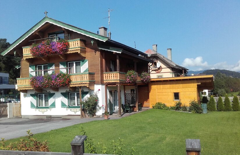 Alpines Wohnhaus mit Holzfassade, Balkonblumen und grün-weißen Fensterläden vor einer Wiese.
