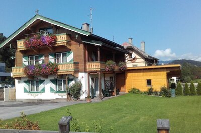 Haus Johanna in den Alpen Alpines Wohnhaus mit Holzfassade, Balkonblumen und grün-weißen Fensterläden vor einer Wiese.
