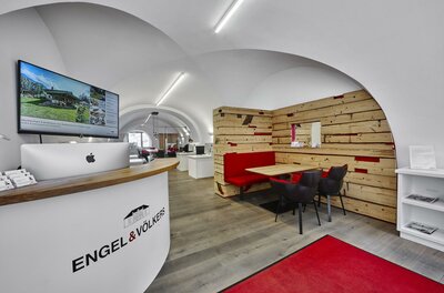 Engel & Völkers Immobilien Ein modernes Büro mit einer Empfangstheke und einem Holzregal. Der Raum ist hell und einladend, mit einem großen Bildschirm und einem roten Teppich.