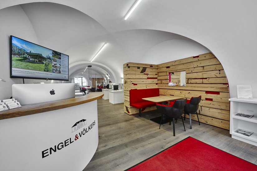 Engel & Völkers Immobilien Ein modernes Büro mit einer Empfangstheke und einem Holzregal. Der Raum ist hell und einladend, mit einem großen Bildschirm und einem roten Teppich.