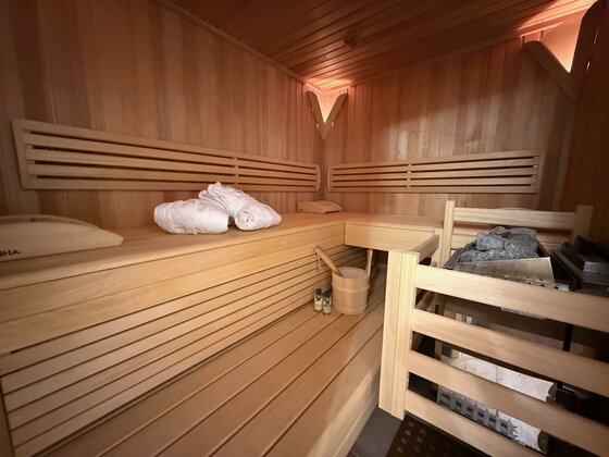 Ein gemütlicher Saunaraum aus Holz mit Sitzbänken und einem Ofen. Handtücher und ein Eimer sind ebenfalls im Raum vorhanden.