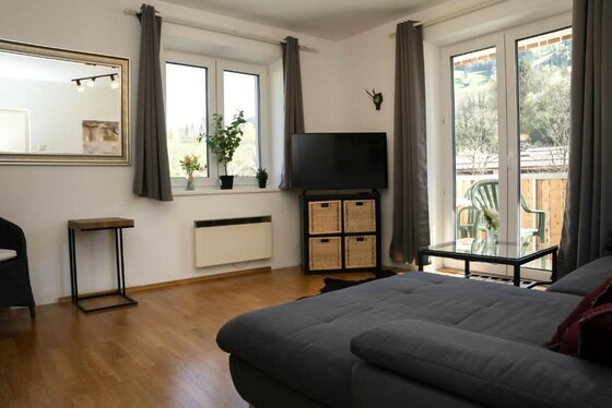 587402278 Ein gemütlicher Wohnraum mit grauem Sofa und Fernseher. Große Fenster bringen viel Tageslicht herein und bieten einen Blick nach draußen.