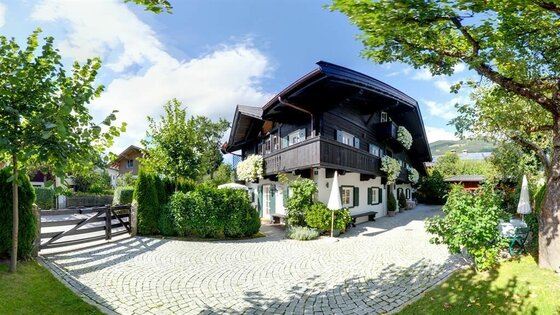 Pano01 Weberhaus Zweistöckiges Alpenhaus mit dunkler Holzfassade, Balkon mit Blumen und gepflastertem Vorhof unter blauem Himmel.