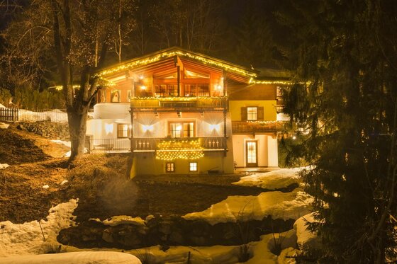 Alpines Ferienhaus bei Nacht Beleuchtetes alpines Holzhaus bei Nacht mit Schneeresten im Vordergrund.