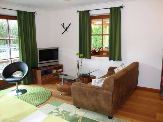 Wohnraum im Haus Andreas Wohnzimmer mit braunem Sofa, Glastisch, Fernseher und grünen Vorhängen vor Holzfenstern.