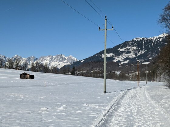 Winterlandschaft bei Kitzbühel Verschneites Feld mit Strommast, kleiner Holzhütte und schneebedeckten Bergen im Hintergrund unter blauem Himmel.