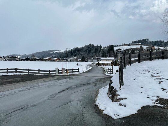 Asphaltierte Straße führt durch eine verschneite Landschaft mit Holzzäunen und Häusern im Hintergrund.