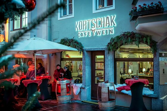Außenansicht des Cafés Kortschak im Winter mit beleuchtetem Schriftzug, Stehtischen und Gästen.