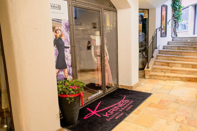 Eingang Krines Style Eingangsbereich eines Modegeschäfts mit Schaufenster, Werbeplakat und einer schwarzen Fußmatte mit pinkem Logo.
