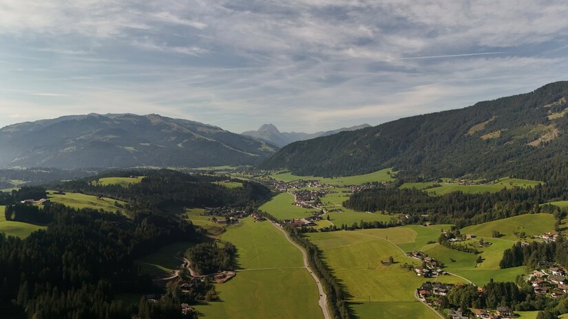 Luftaufnahme eines Tals mit grünen Wiesen, kleinen Siedlungen und bewaldeten Bergen unter leicht bewölktem Himmel.