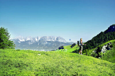 Wanderin mit Rucksack auf einer grünen Bergwiese vor einem Gebirgspanorama mit Seilbahngondel.